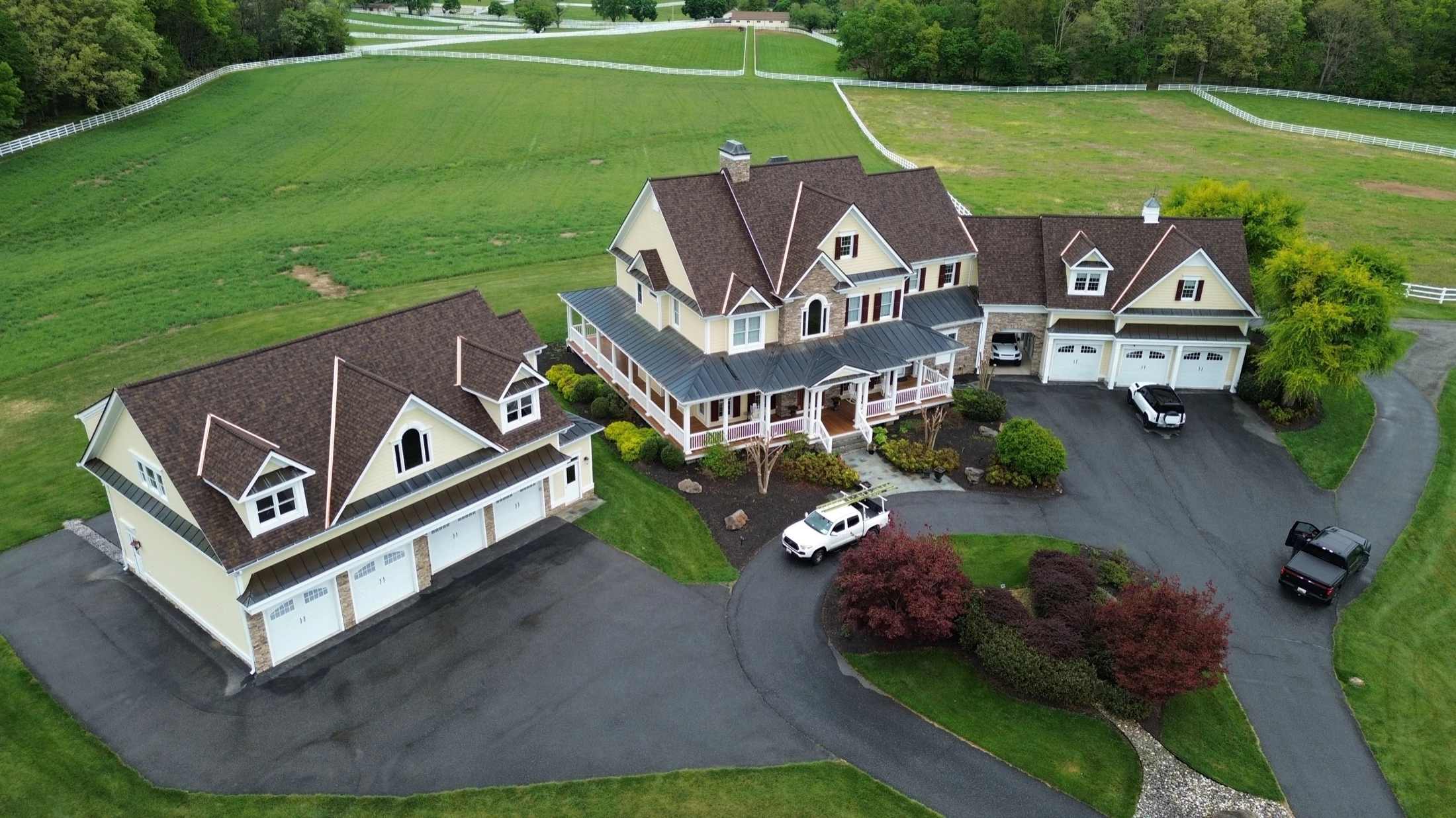 Aerial drone view of a completed Supreme Restorations residential roof on a Maryland estate property, brown architectural asphalt shingles installed over a multi-section home with attached garage and white horse fencing across the surrounding pasture